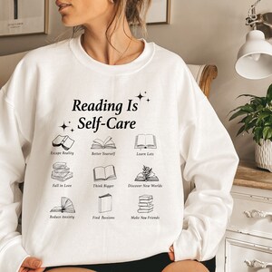May include: White sweatshirt with the text "Reading Is Self-Care" and illustrations of books with phrases like "Escape Reality", "Better Yourself", "Learn Lots", "Fell in Love", "Think Bigger", "Discover New Worlds", "Reduce Anxiety", "Find Passions", and "Make New Friends".