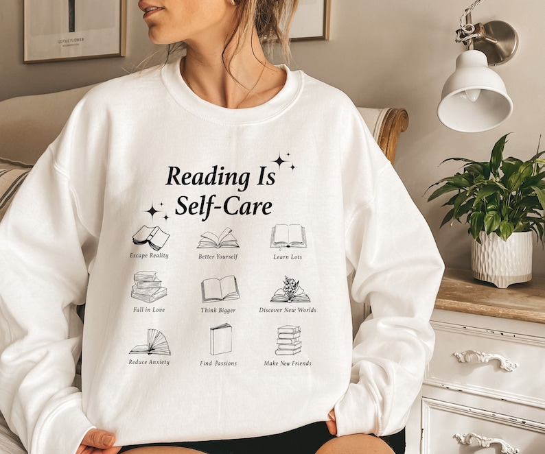 May include: White sweatshirt with the text "Reading Is Self-Care" and illustrations of books with phrases like "Escape Reality", "Better Yourself", "Learn Lots", "Fell in Love", "Think Bigger", "Discover New Worlds", "Reduce Anxiety", "Find Passions", and "Make New Friends".