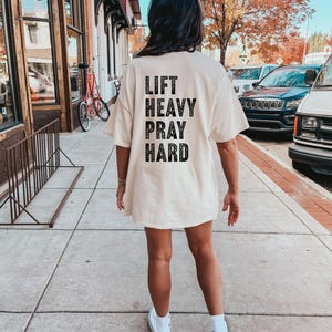May include: A white oversized t-shirt with black text that reads "Lift Heavy Pray Hard".