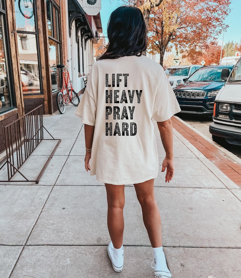 May include: A white oversized t-shirt with black text that reads "Lift Heavy Pray Hard".