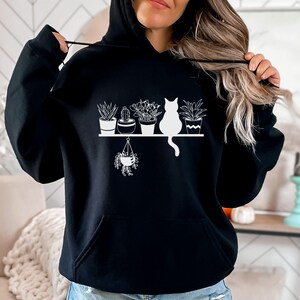May include: Black hoodie with a white graphic of a cat sitting on a shelf with potted plants. The graphic includes a hanging plant.