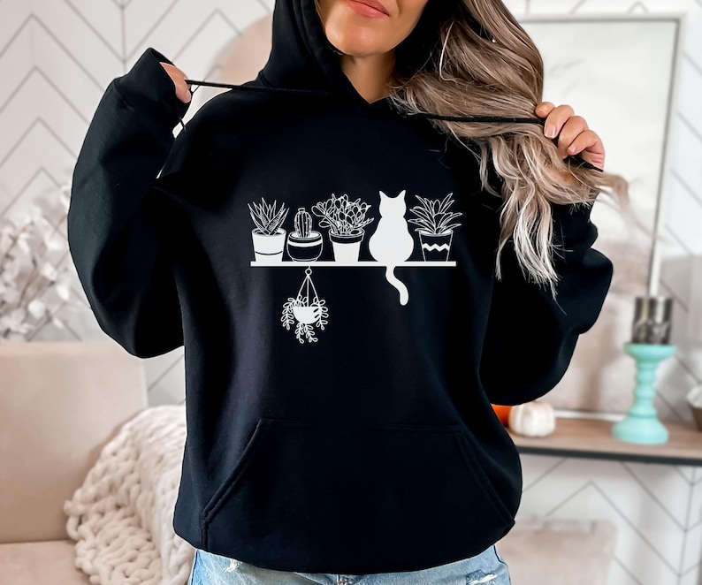 May include: Black hoodie with a white graphic of a cat sitting on a shelf with potted plants. The graphic includes a hanging plant.