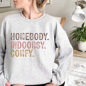 以下が含まれることがあります： グレーのトレーナーに「HOMEBODY. INDOORSY. COMFY.」という文字が茶色、ピンク、金色の文字で書かれています。