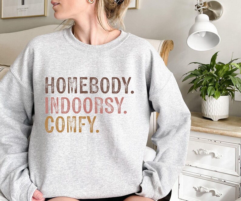 以下が含まれることがあります： グレーのトレーナーに「HOMEBODY. INDOORSY. COMFY.」という文字が茶色、ピンク、金色の文字で書かれています。