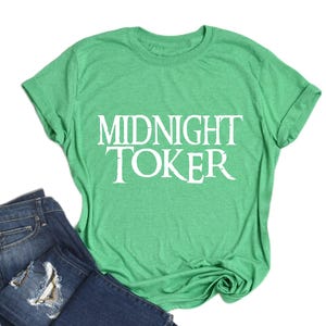 Puede incluir: Una camiseta verde con el texto blanco "MIDNIGHT TOKER", junto con un par de vaqueros azules. La camiseta es de color verde liso, y los vaqueros tienen un aspecto desgastado.