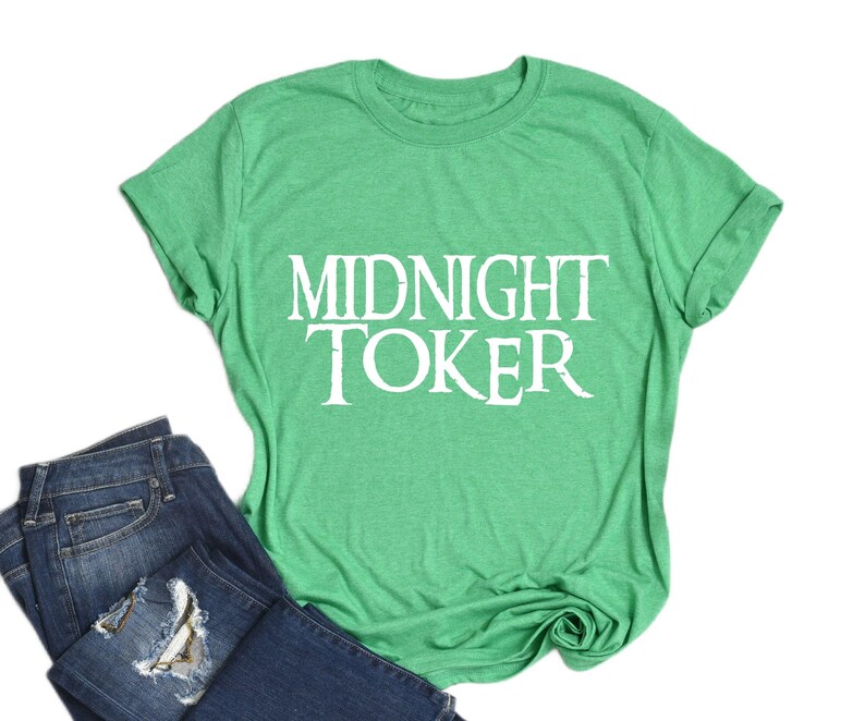 Puede incluir: Una camiseta verde con el texto blanco "MIDNIGHT TOKER", junto con un par de vaqueros azules. La camiseta es de color verde liso, y los vaqueros tienen un aspecto desgastado.