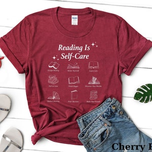 May include: A cherry red t-shirt with the text "Reading Is Self-Care" and illustrations of books with phrases like "Escape Reality", "Better Yourself", "Learn Lots", "Fall in Love", "Think Bigger", "Discover New Worlds", "Reduce Anxiety", "Find Passions", and "Make New Friends".