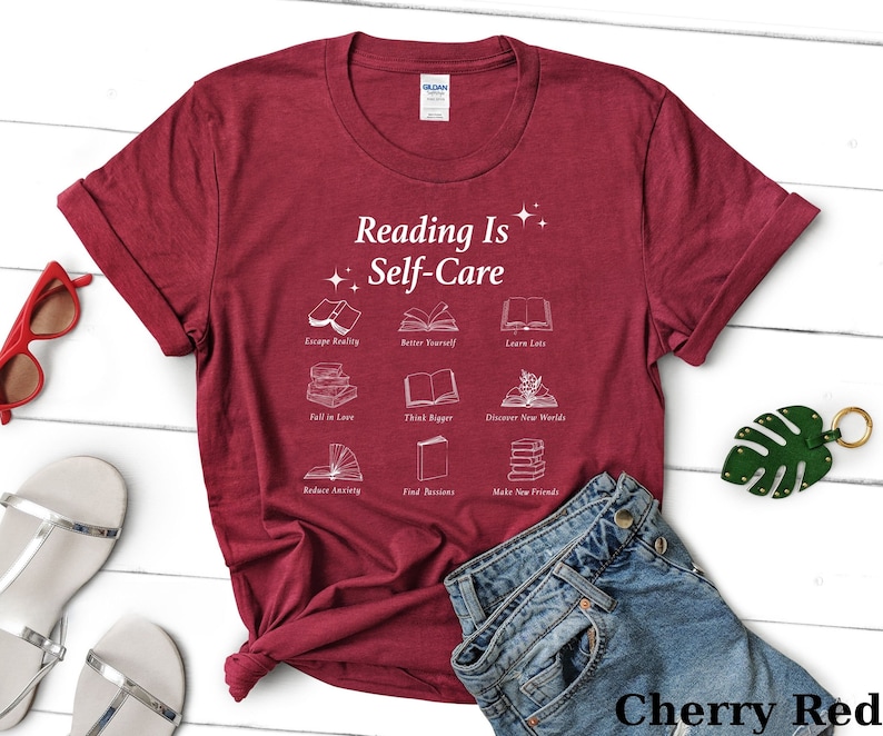 May include: A cherry red t-shirt with the text "Reading Is Self-Care" and illustrations of books with phrases like "Escape Reality", "Better Yourself", "Learn Lots", "Fall in Love", "Think Bigger", "Discover New Worlds", "Reduce Anxiety", "Find Passions", and "Make New Friends".