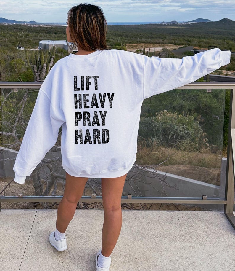 May include: White long-sleeve sweatshirt with the text "LIFT HEAVY PRAY HARD" printed on the back in black.