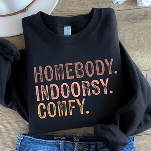 以下が含まれることがあります： オレンジ、ピンク、イエローの色で、くたびれたフォントで「Homebody. Indoorsy. Comfy.」という文字が書かれた黒いスウェットシャツです。