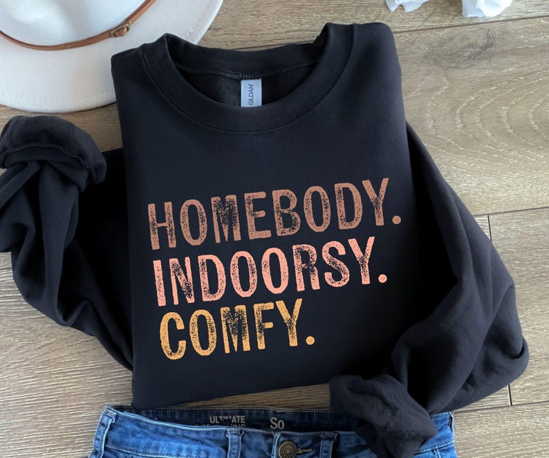 以下が含まれることがあります： オレンジ、ピンク、イエローの色で、くたびれたフォントで「Homebody. Indoorsy. Comfy.」という文字が書かれた黒いスウェットシャツです。