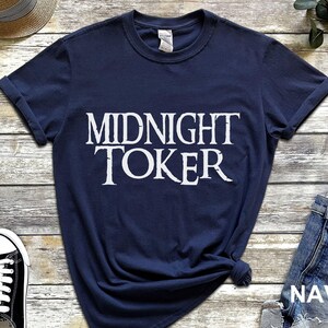 Puede incluir: Camiseta azul marino con la frase "MIDNIGHT TOKER" en blanco. La camiseta tiene las mangas dobladas y un nudo en la parte inferior. Tambi&eacute;n se ven zapatillas deportivas blancas y negras y unos pantalones cortos vaqueros.