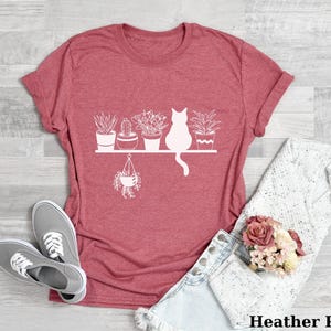 May include: A heather pink t-shirt with a white graphic of a cat sitting on a shelf with potted plants. The text "Heather Pink" is at the bottom of the image.