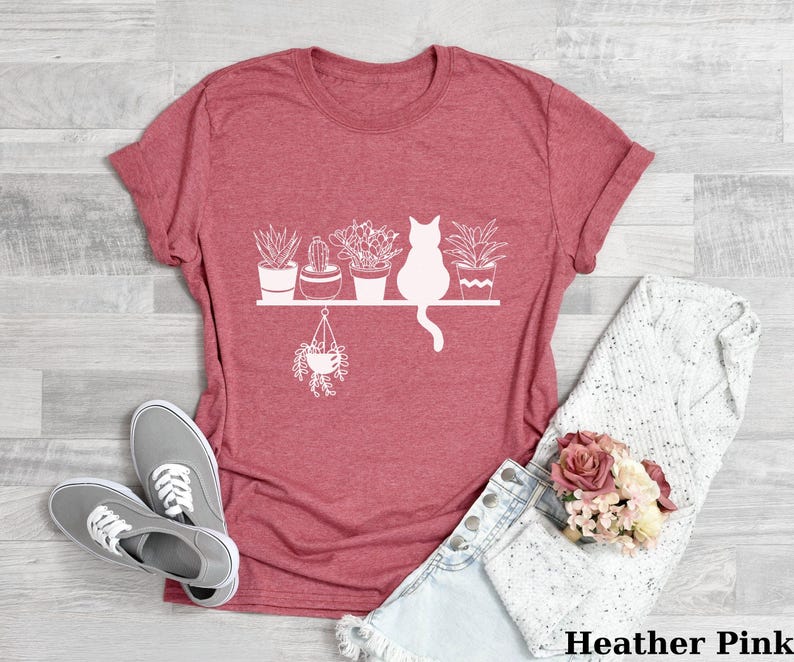 May include: A heather pink t-shirt with a white graphic of a cat sitting on a shelf with potted plants. The text "Heather Pink" is at the bottom of the image.