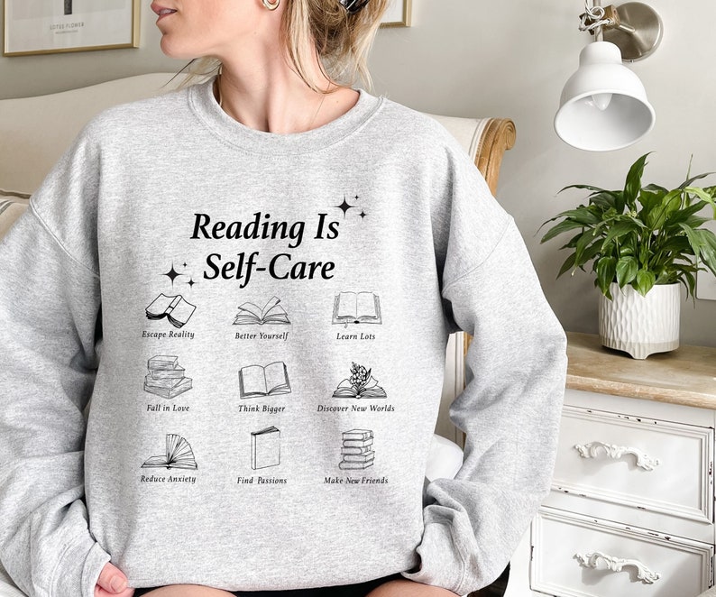 May include: Gray sweatshirt with the text "Reading Is Self-Care" and illustrations of books with phrases like "Escape Reality", "Better Yourself", "Learn Lots", "Fall in Love", "Think Bigger", "Discover New Worlds", "Reduce Anxiety", "Find Passions", and "Make New Friends".