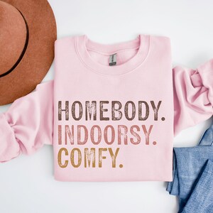 以下が含まれることがあります： ピンクのトレーナーに「Homebody. Indoorsy. Comfy.」という文字が茶色、オレンジ、金色の文字で書かれています。