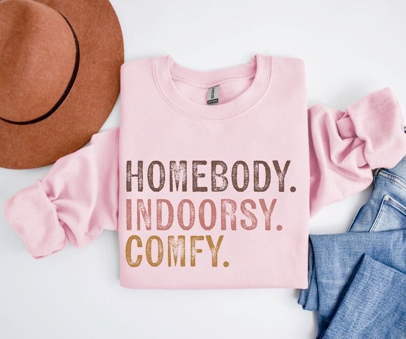 以下が含まれることがあります： ピンクのトレーナーに「Homebody. Indoorsy. Comfy.」という文字が茶色、オレンジ、金色の文字で書かれています。