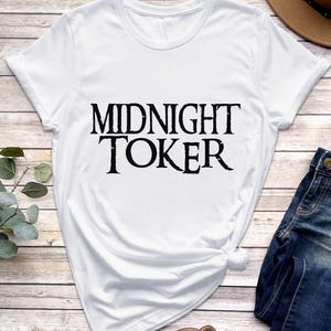 Puede incluir: Camiseta blanca con el texto "MIDNIGHT TOKER" en una fuente negra audaz y desgastada. La camiseta se combina con vaqueros azules y un sombrero marr&oacute;n, sobre un fondo de vetas de madera clara.