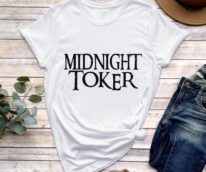 Puede incluir: Camiseta blanca con el texto "MIDNIGHT TOKER" en una fuente negra audaz y desgastada. La camiseta se combina con vaqueros azules y un sombrero marr&oacute;n, sobre un fondo de vetas de madera clara.