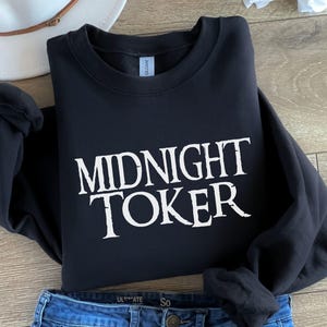 Puede incluir: Sudadera negra con el texto blanco "MIDNIGHT TOKER" en una fuente audaz y estilizada. La sudadera est&aacute; doblada, con unos vaqueros azules y un sombrero blanco en el fondo.