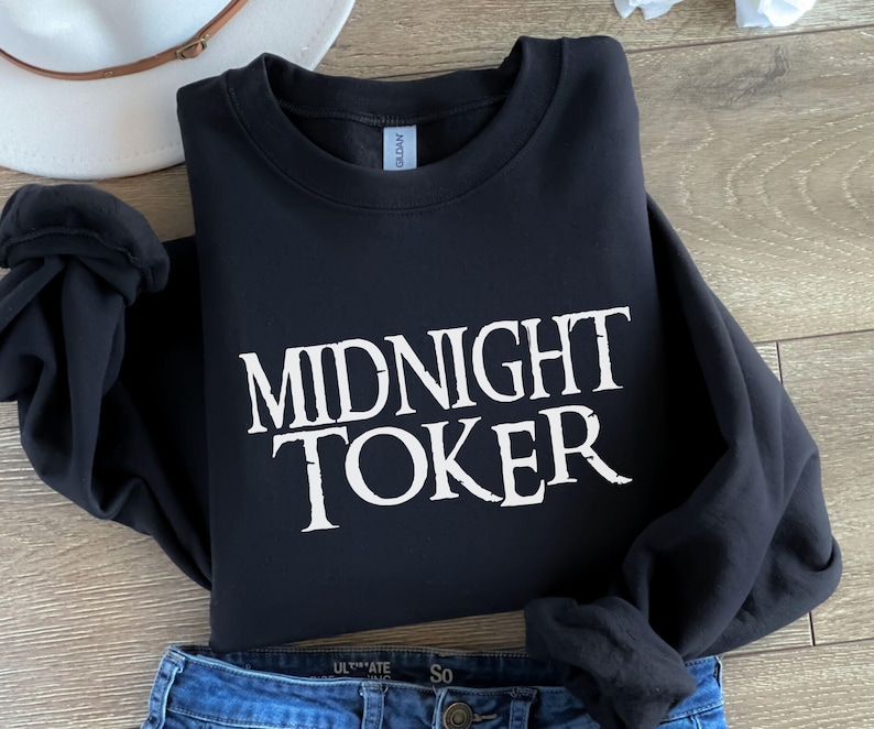 Puede incluir: Sudadera negra con el texto blanco "MIDNIGHT TOKER" en una fuente audaz y estilizada. La sudadera est&aacute; doblada, con unos vaqueros azules y un sombrero blanco en el fondo.