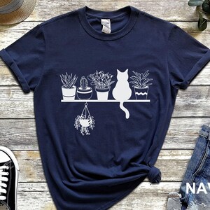 May include: Navy blue t-shirt with a white graphic of a cat sitting in front of a shelf with potted plants. The text "NAVY" is printed in white on the bottom right corner of the image.