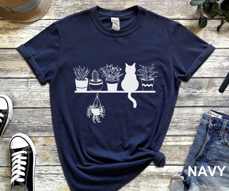 May include: Navy blue t-shirt with a white graphic of a cat sitting in front of a shelf with potted plants. The text "NAVY" is printed in white on the bottom right corner of the image.