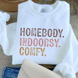 以下が含まれることがあります： ブラウン、ピンク、オレンジで「Homebody. Indoorsy. Comfy.」とプリントされた白いスウェットシャツです。