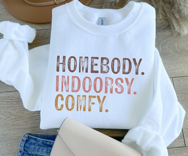 以下が含まれることがあります： ブラウン、ピンク、オレンジで「Homebody. Indoorsy. Comfy.」とプリントされた白いスウェットシャツです。