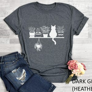 May include: Dark gray heather t-shirt with a white graphic of a cat sitting on a shelf with potted plants. The text "DARK GRAY (HEATHER)" is printed on the bottom right corner of the image.