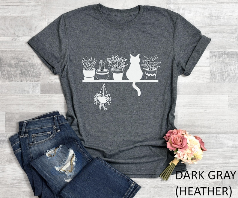 May include: Dark gray heather t-shirt with a white graphic of a cat sitting on a shelf with potted plants. The text "DARK GRAY (HEATHER)" is printed on the bottom right corner of the image.