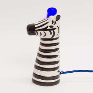 May include: A ceramic zebra-shaped lamp with a black and white striped design. The lamp emits a blue light from the top. A blue cord is attached to the base of the lamp.