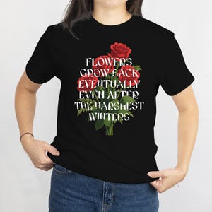 May include: Black t-shirt featuring a floral design with red roses and the text "FLOWERS GROW BACK EVENTUALLY EVEN AFTER THE HARSHEST WINTERS" in white. The shirt is a crew neck and made of a soft material.