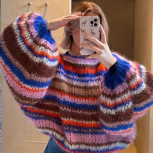 Puede incluir: Un suéter de punto de rayas de colores con una textura esponjosa. El suéter tiene un corte holgado y mangas largas con un puño ancho y fruncido.