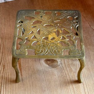 May include: A square brass trivet featuring an intricate openwork design with a central crest, crossed swords, and the text "DIEU ET MON DROIT" embossed along the bottom edge, supported by four curved legs with a polished metallic finish.