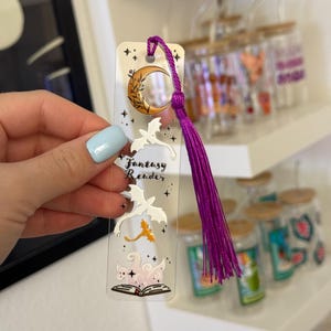 Könnte beinhalten: Ein transparentes Acryl-Lesezeichen mit einem violetten Quasten. Das Lesezeichen zeigt einen Halbmond, Sterne, ein Buch und zwei Drachen. Der Text "Fantasy Reader" ist auf dem Lesezeichen gedruckt.