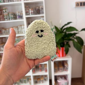 Op de afbeelding: Een lichtgroene gehaakte spons in de vorm van een spookje met een textuur. Het spookje heeft geborduurde zwarte stervormige ogen, een zwarte gebogen mond en roze wangen. Een klein label met de tekst Plants Coffee is aan de zijkant bevestigd.