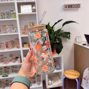 Könnte beinhalten: Ein klarer Glastrinkbecher mit Bambusdeckel und Glasstrohhalm, verziert mit einem bunten Blumenmuster aus orangefarbenen, roten und hellrosa Gänseblümchen sowie grünen Blättern, die auf die Außenseite des zylindrischen Gefäßes gedruckt sind.