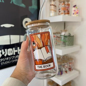 Könnte beinhalten: Ein klares Glasgefäß mit Bambusdeckel und Strohhalm. Das Gefäß ist mit einer bunten Illustration eines geöffneten Buches, einer Tasse Kaffee und dem Text "THE BOOK" verziert.