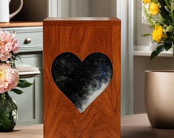 Puede incluir: Una urna rectangular de madera con un acabado de grano marrón cálido que presenta un gran recorte en forma de corazón en el panel frontal. El recorte revela una superficie interior texturizada, veteada en negro y blanco, que contrasta.