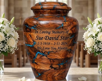 May include: A wooden urn with a lid, featuring a brown and blue design. The urn has text that reads "In Loving Memory of Sue David Star 2-3-1958 - 2-11-2024 We Love You Always". White flowers in vases flank the urn.