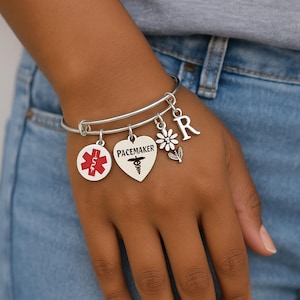 Puede incluir: Pulsera de plata con múltiples colgantes. Los colgantes incluyen un símbolo médico rojo y blanco, un colgante en forma de corazón con la palabra "Pacemaker" y un símbolo de caduceo, una margarita y la letra "R".
