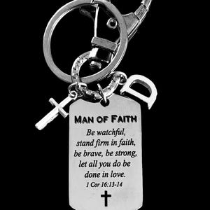 May include: Silver-toned keychain with a dog tag pendant. The tag reads "MAN OF FAITH" and includes an inspirational quote and a cross symbol. A small cross charm and a letter "D" charm are also attached.