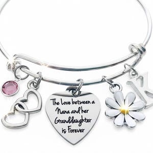 May include: Silver bangle bracelet with various charms. Charms include a pink gemstone, two interlocking hearts, a heart-shaped pendant with text, a white daisy, and a letter "K". The heart pendant reads "The Love between a Nana and her Granddaughter is Forever".