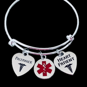 Könnte beinhalten: Ein silbernes Armreif-Armband mit drei Anhängern. Zwei herzförmige Anhänger tragen die Aufschrift "Pacemaker" und "Heart Patient" mit medizinischen Symbolen. Ein runder Anhänger zeigt ein rotes Medizinsymbol. Das Armband ist vor schwarzem Hintergrund.