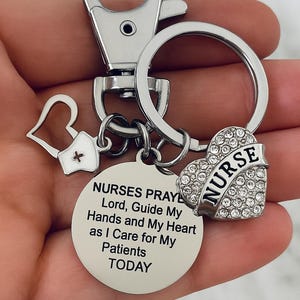 May include: Silver-colored keychain with a heart-shaped charm encrusted with rhinestones and the word "NURSE". Includes a round charm with a prayer for nurses. The keychain also has a small heart and a medical cross charm. The text "NURSES PRAYER Keychain" is at the bottom.