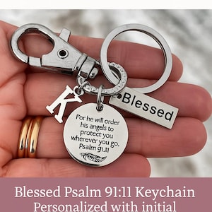 May include: A silver-coloured keychain with a carabiner clip, a ring, and a charm that says "Blessed." It includes a round charm with a Bible verse and a letter "K" charm. The keychain is a faith-based accessory.
