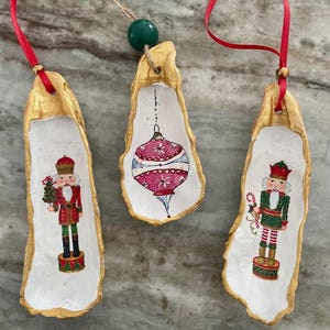 Peut inclure: Trois décorations de Noël faites à partir de coquilles d'huîtres. Chaque coquille est peinte avec un bord doré et un intérieur blanc. Les ornements présentent des motifs festifs, dont un casse-noisette et une boule de Noël. Des rubans rouges sont attachés pour les suspendre.