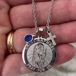 Op de afbeelding: Zilverkleurige ketting met een hanger met de afbeelding van Sint-Raphaël, de tekst "Saint Raphael" en "Pray for Us". De ketting bevat ook een blauwe edelsteen en een kleine kruisbedel.