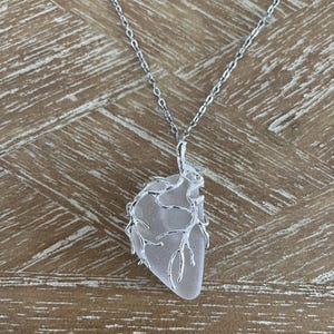May include: A silver-toned necklace featuring a pendant shaped like an anatomical heart. The pendant consists of a translucent, frosted white stone encased in a silver-colored metal frame that mimics the appearance of branching veins and arteries.